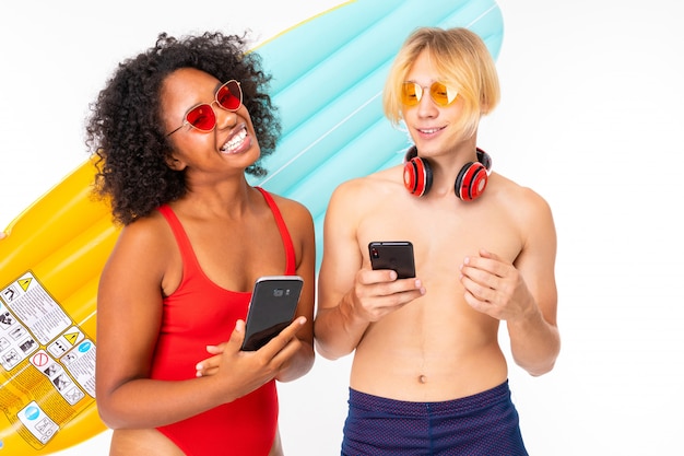 Il giovane uomo biondo femminile e caucasico sta in costume da bagno con i grandi materassi di gomma della spiaggia, comunica con gli amici sui telefoni e sui sorrisi, isolati su bianco