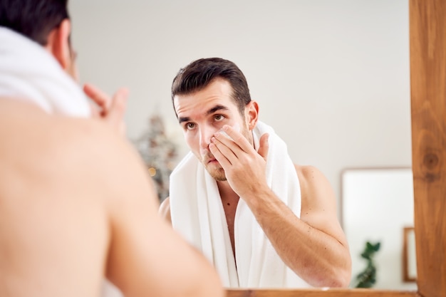 Il giovane uomo brunet spalma la crema sul viso mentre si trova vicino allo specchio nella vasca da bagno al mattino