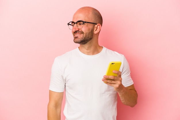 Il giovane uomo calvo caucasico che tiene il telefono cellulare isolato su sfondo rosa sembra da parte sorridente, allegro e piacevole.