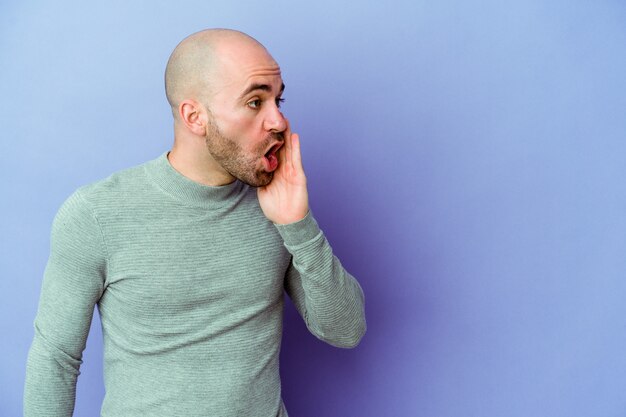 Il giovane uomo calvo caucasico isolato su sfondo viola sta dicendo una notizia segreta di frenata calda e sta guardando da parte