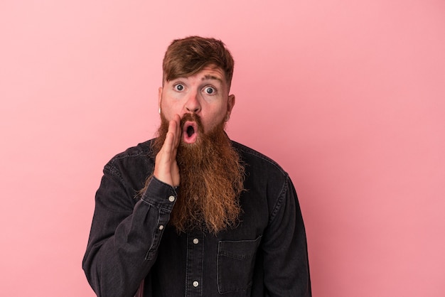 Il giovane uomo caucasico allo zenzero con la barba lunga isolato su sfondo rosa sta dicendo una notizia segreta di frenata calda e sta guardando da parte