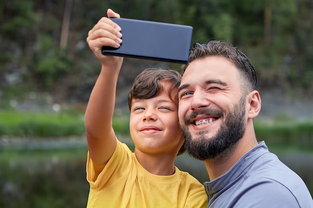 Il giovane uomo caucasico barbuto e suo figlio stanno sorridendo mentre guardano la macchina fotografica sul telefono cellulare