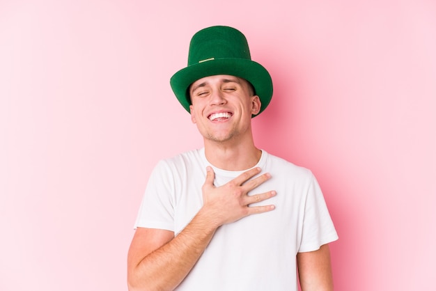 Il giovane uomo caucasico che celebra il giorno di San Patrizio ride fragorosamente tenendo la mano sul petto.