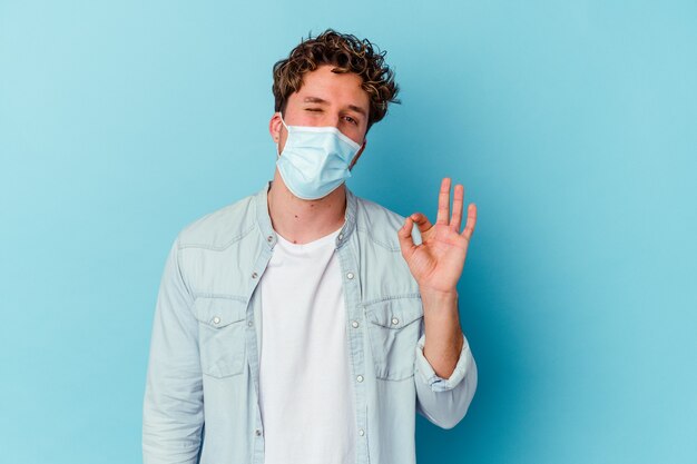 Il giovane uomo caucasico che indossa una maschera antivirale isolata su sfondo blu strizza l'occhio e tiene un gesto ok con la mano.