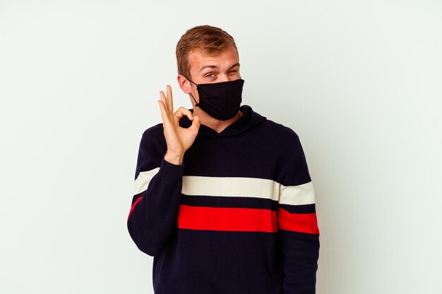 Il giovane uomo caucasico che indossa una maschera per il virus isolato su bianco strizza l'occhio e tiene un gesto giusto con la mano.