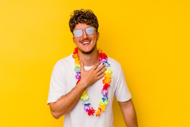 Il giovane uomo caucasico che indossa una roba da festa hawaiana isolata sul muro giallo ride ad alta voce tenendo la mano sul petto.