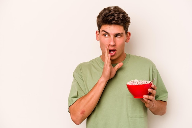 Il giovane uomo caucasico che mangia cereali isolati su sfondo bianco sta dicendo una notizia segreta di frenata calda e sta guardando da parte
