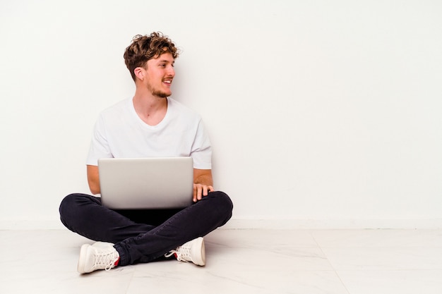 Il giovane uomo caucasico che si siede sul pavimento che tiene sul computer portatile isolato su fondo bianco guarda da parte sorridente, allegro e piacevole.