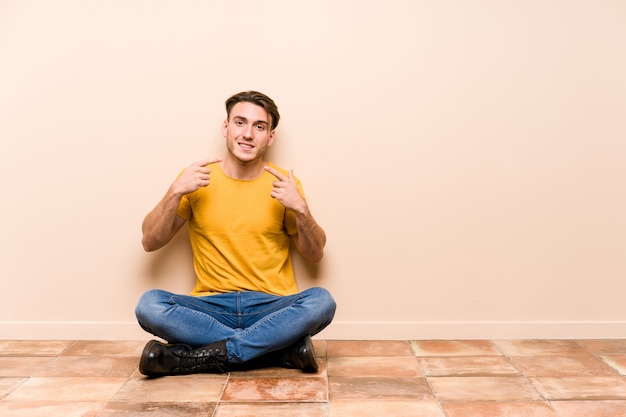 Il giovane uomo caucasico che si siede sul pavimento ha isolato i sorrisi, indicando le dita alla bocca.