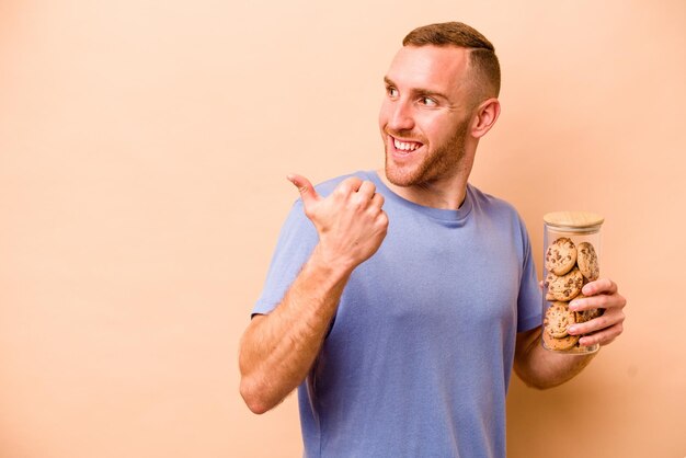 Il giovane uomo caucasico che tiene il barattolo di biscotti isolato su sfondo beige indica con il dito del pollice lontano ridendo e spensierato