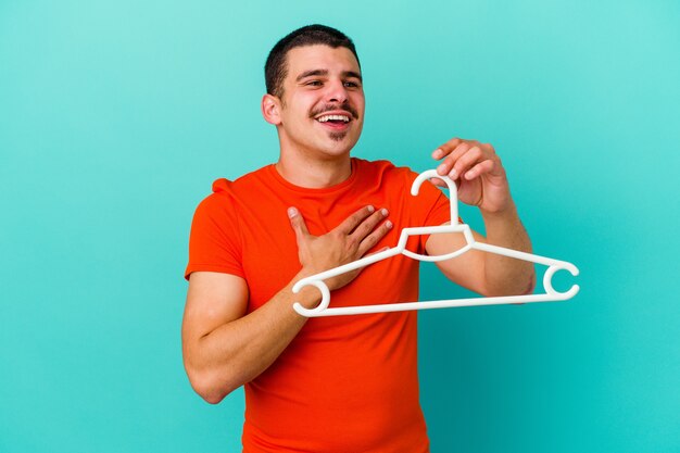 Il giovane uomo caucasico che tiene un gancio isolato sulla parete blu ride ad alta voce mantenendo la mano sul petto.