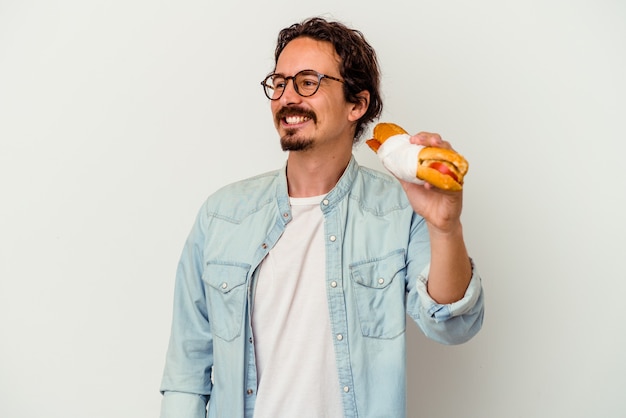 Il giovane uomo caucasico che tiene un panino isolato su bianco guarda da parte sorridente, allegro e piacevole.