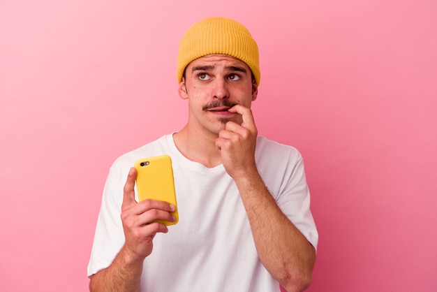 Il giovane uomo caucasico che tiene un telefono cellulare isolato su sfondo rosa si è rilassato pensando a qualcosa guardando uno spazio di copia.