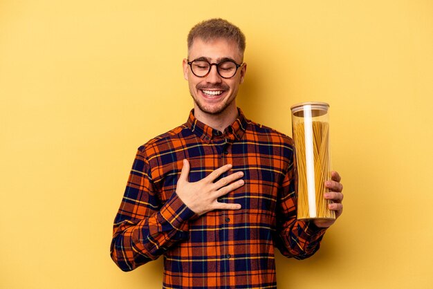Il giovane uomo caucasico che tiene un vasetto di spaghetti isolato su sfondo giallo ride ad alta voce tenendo la mano sul petto.