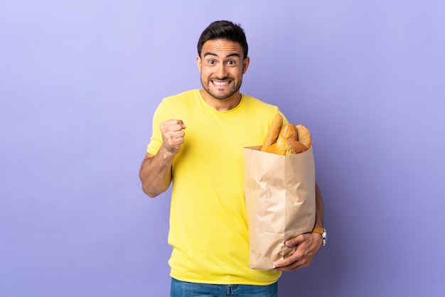 Il giovane uomo caucasico che tiene una borsa piena di pani ha isolato che celebra una vittoria