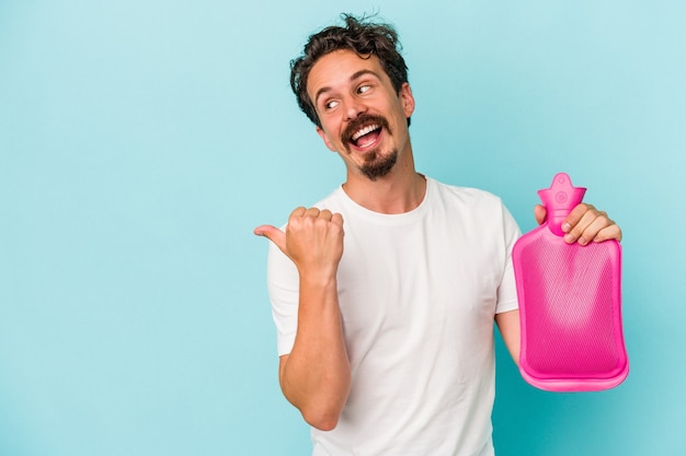 Il giovane uomo caucasico che tiene una sacca d'acqua isolata su sfondo blu punta con il pollice lontano, ridendo e spensierato.