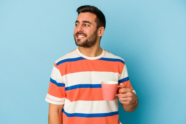Il giovane uomo caucasico che tiene una tazza isolata sulla parete blu guarda da parte sorridente, allegro e piacevole.