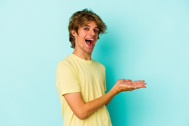 Il giovane uomo caucasico con compone isolato su fondo blu che tiene uno spazio della copia su una palma.