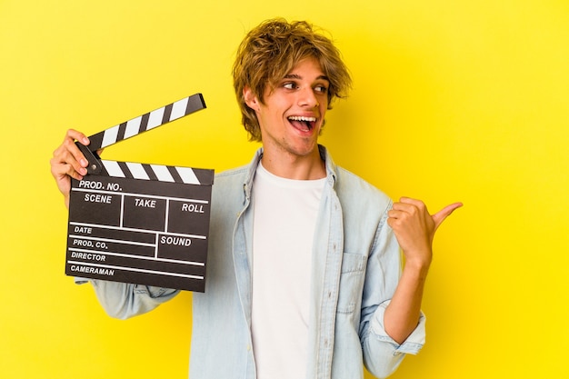 Il giovane uomo caucasico con il ciak della tenuta del trucco isolato su sfondo giallo indica con il dito del pollice lontano, ridendo e spensierato.