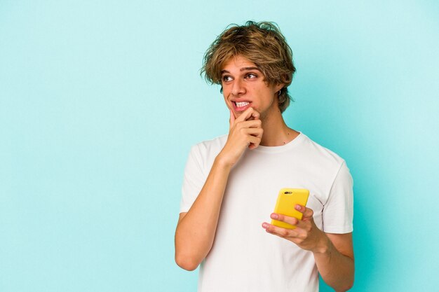 Il giovane uomo caucasico con il trucco che tiene il telefono cellulare isolato su sfondo blu si è rilassato pensando a qualcosa guardando uno spazio di copia.