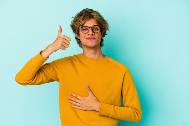 Il giovane uomo caucasico con trucco isolato su sfondo blu tocca la pancia, sorride dolcemente, mangia e soddisfa il concetto.