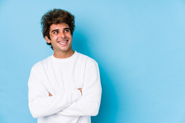 Il giovane uomo caucasico contro una parete blu ha isolato sorridere sicuro con le braccia attraversate.