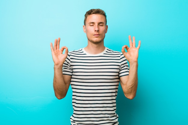 Il giovane uomo caucasico contro una parete blu si rilassa dopo una dura giornata di lavoro, sta eseguendo yoga.