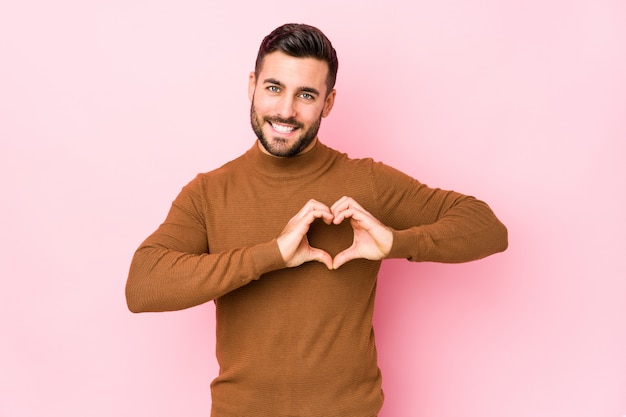 Il giovane uomo caucasico contro una parete rosa ha isolato sorridere e mostrare una forma del cuore con le mani.