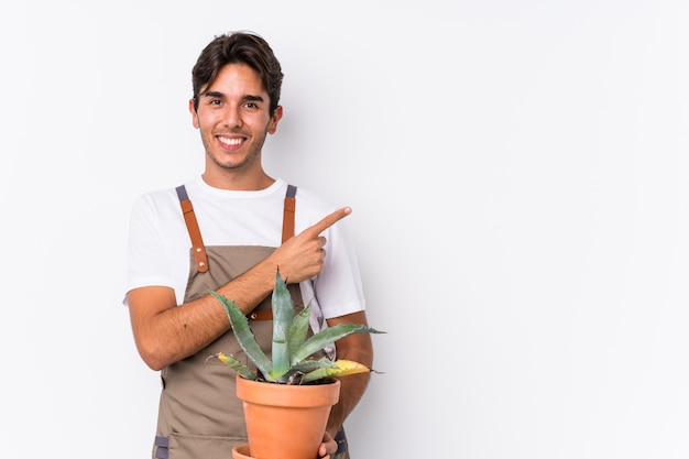 Il giovane uomo caucasico del giardiniere che tiene una pianta ha isolato sorridere e indicare da parte, mostrando qualcosa nello spazio.