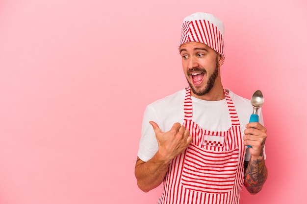 Il giovane uomo caucasico del produttore di ghiaccio con i tatuaggi che tiene il cucchiaio isolato su sfondo rosa indica con il pollice lontano, ridendo e spensierato.