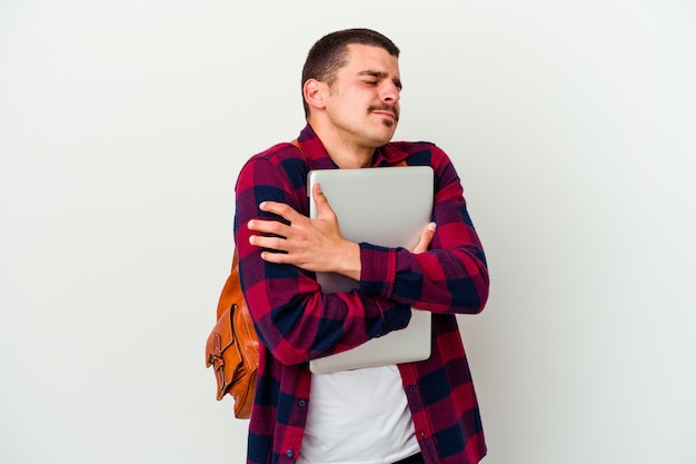 Il giovane uomo caucasico dello studente che tiene un computer portatile isolato sugli abbracci bianchi del fondo, sorride spensierato e felice.