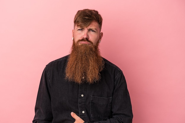 Il giovane uomo caucasico dello zenzero con la barba lunga isolata su fondo rosa tocca la pancia, sorride dolcemente, mangia e concetto di soddisfazione.