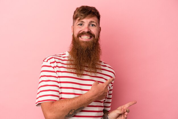 Il giovane uomo caucasico dello zenzero con la barba lunga isolato su fondo rosa ha eccitato indicando con gli indici di distanza.