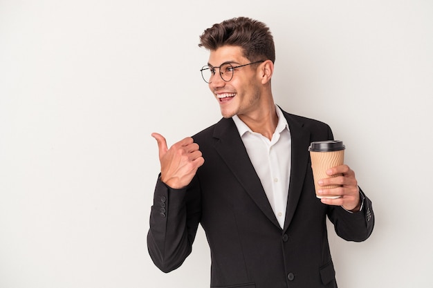 Il giovane uomo caucasico di affari che tiene caffè da asporto isolato su sfondo bianco indica con il dito pollice lontano, ridendo e spensierato.