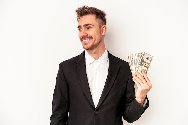 Il giovane uomo caucasico di affari che tiene le banconote isolate su fondo bianco guarda da parte sorridente, allegro e piacevole.