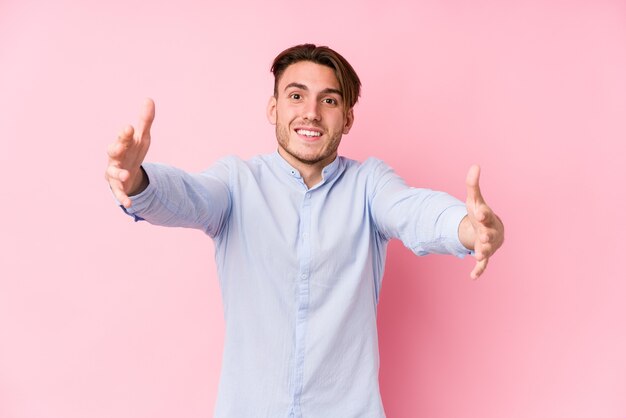 Il giovane uomo caucasico in posa in uno sfondo rosa isolato si sente sicuro di dare un abbraccio alla telecamera.