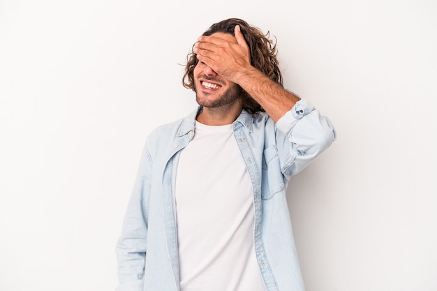 Il giovane uomo caucasico isolato su sfondo bianco copre gli occhi con le mani, sorride ampiamente in attesa di una sorpresa.