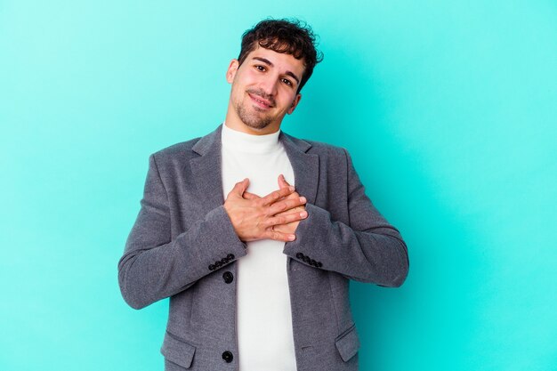 Il giovane uomo caucasico isolato su sfondo blu ha un'espressione amichevole, premendo il palmo sul petto. Concetto di amore.