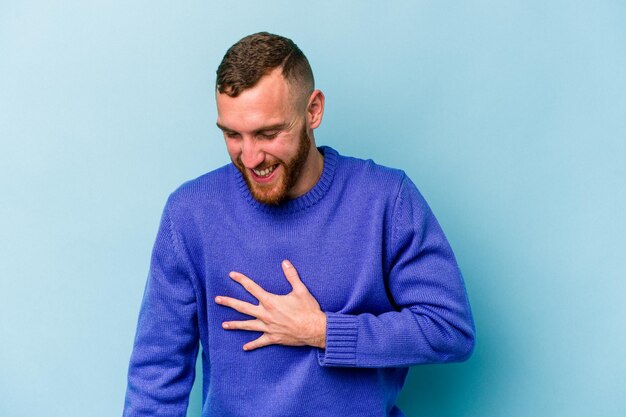 Il giovane uomo caucasico isolato su sfondo blu ride ad alta voce tenendo la mano sul petto.