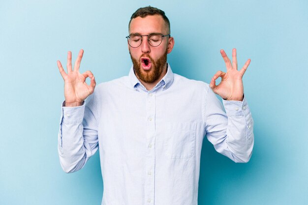 Il giovane uomo caucasico isolato su sfondo blu si rilassa dopo una dura giornata di lavoro, sta eseguendo yoga.