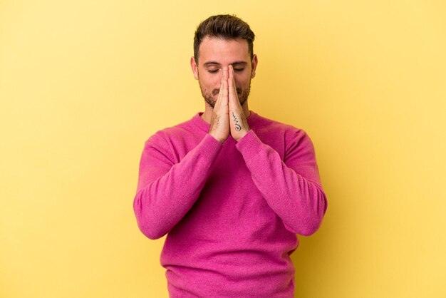 Il giovane uomo caucasico isolato su sfondo giallo che si tiene per mano in preghiera vicino alla bocca, si sente sicuro.