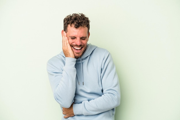 Il giovane uomo caucasico isolato su sfondo verde ride felice e si diverte a tenere le mani sullo stomaco.