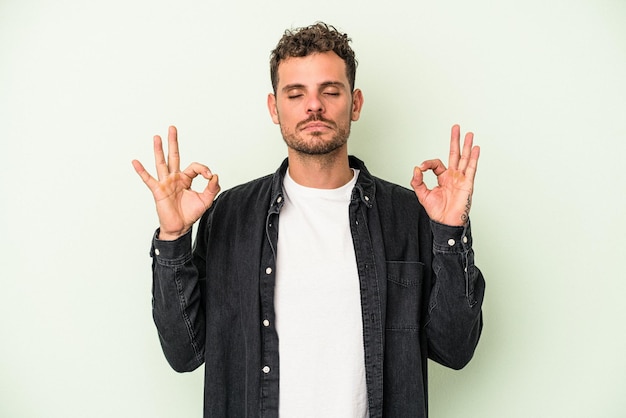 Il giovane uomo caucasico isolato su sfondo verde si rilassa dopo una dura giornata di lavoro, sta eseguendo yoga.