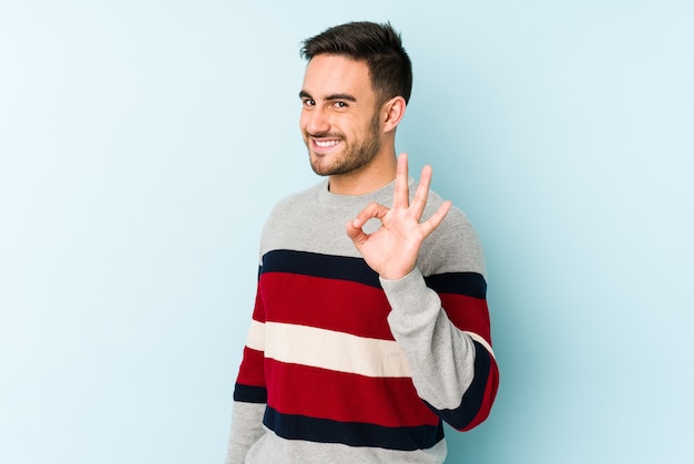 Il giovane uomo caucasico isolato sull'azzurro fa l'occhiolino di un occhio e tiene un gesto giusto con la mano.