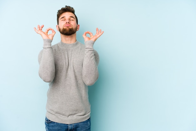 Il giovane uomo caucasico isolato sulla parete blu si rilassa dopo una dura giornata di lavoro, sta eseguendo yoga.