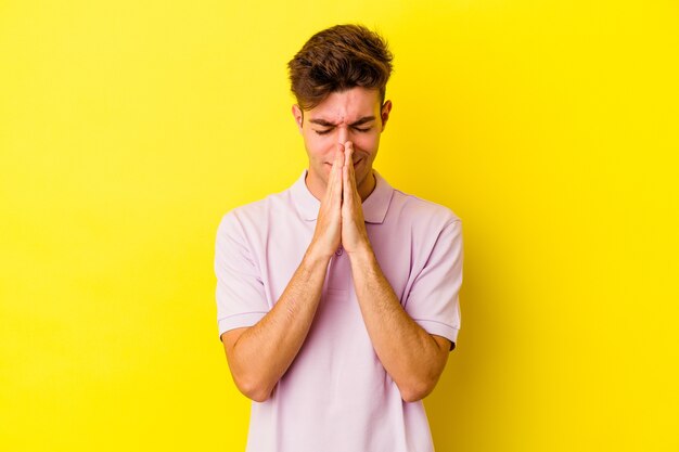 Il giovane uomo caucasico isolato sulla parete gialla che tiene le mani nella preghiera vicino alla bocca, si sente sicuro.