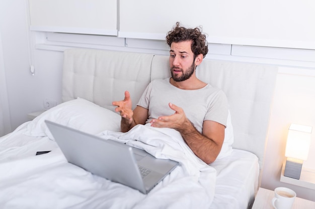 Il giovane uomo caucasico prende il caffè mattutino, si siede sul letto parlando con una videochiamata, legge un libro, si prepara per l'esame, usa il computer portatile per navigare in Internet, vestito con abiti domestici casual.