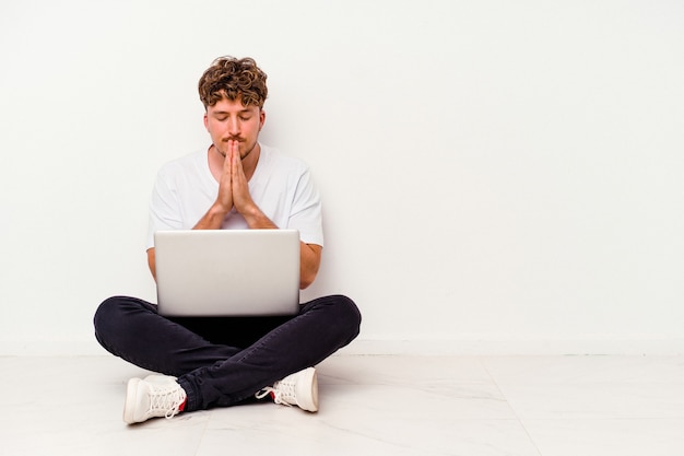 Il giovane uomo caucasico seduto sul pavimento che tiene su un laptop isolato su sfondo bianco tenendosi per mano in preghiera vicino alla bocca, si sente sicuro.