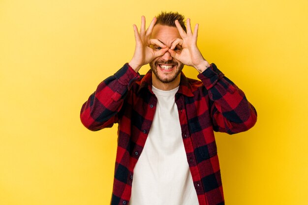 Il giovane uomo caucasico tatuato isolato sulla parete gialla eccitato mantenendo il gesto giusto sull'occhio.