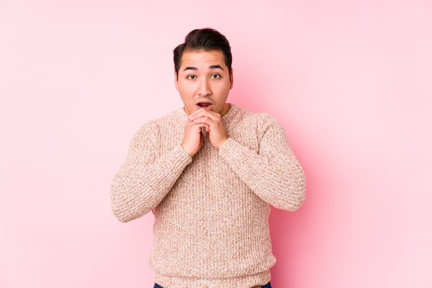 Il giovane uomo curvy che posa in una parete rosa ha isolato pregare per fortuna, bocca stupita e d'apertura che guarda alla parte anteriore.